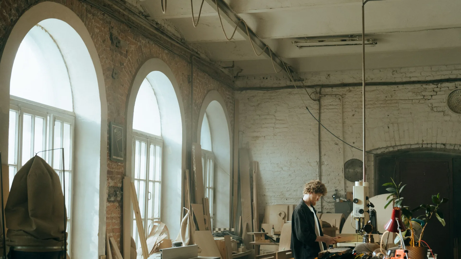 Un grand atelier de brique aux fenêtres en arc : de la lumière du jour, du bois débité partout, un menuisier au fond à l'établi.