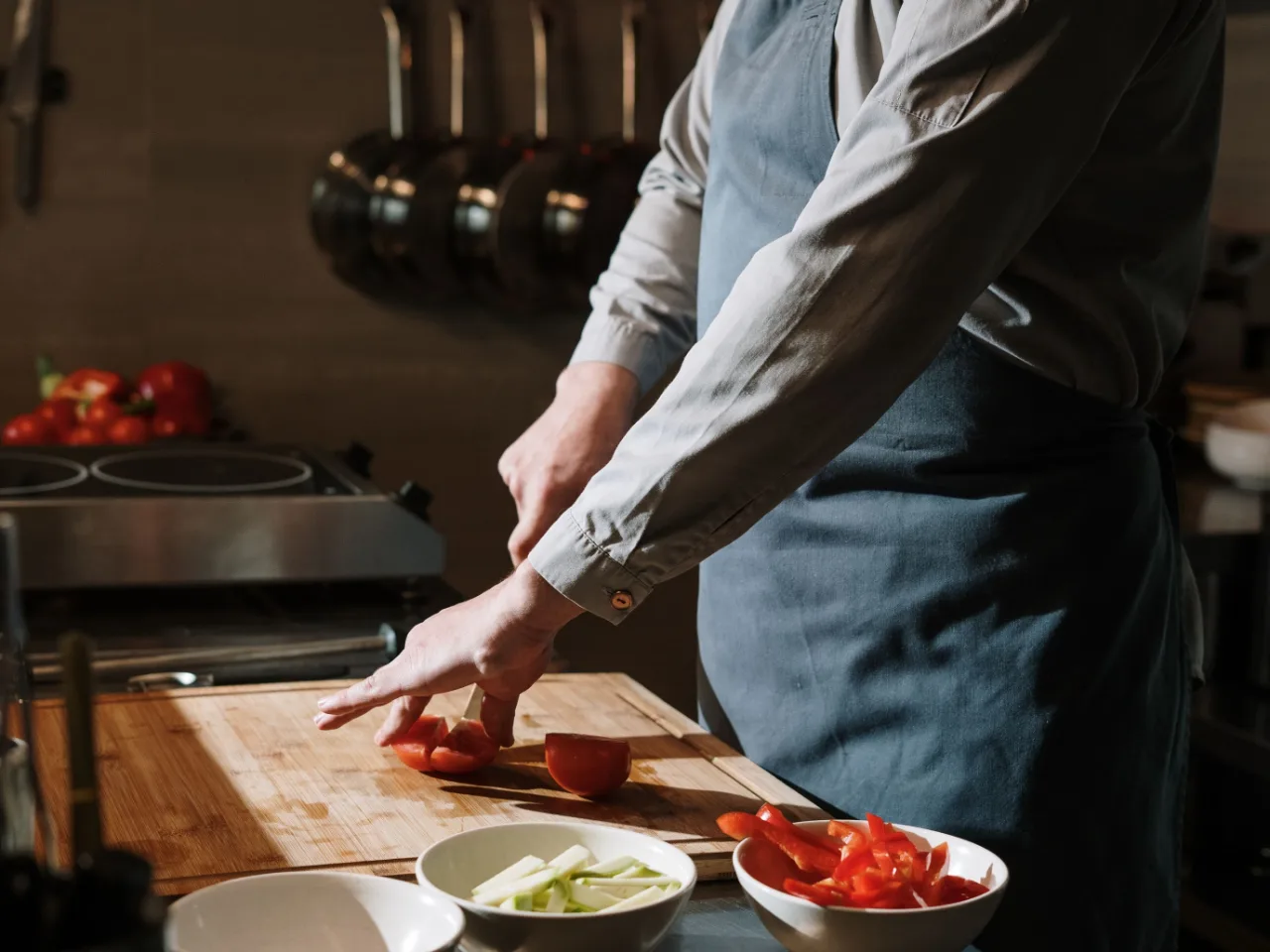 Des mains taillent une tomate dans une cuisine de restaurant, cuivres suspendus au-dessus.