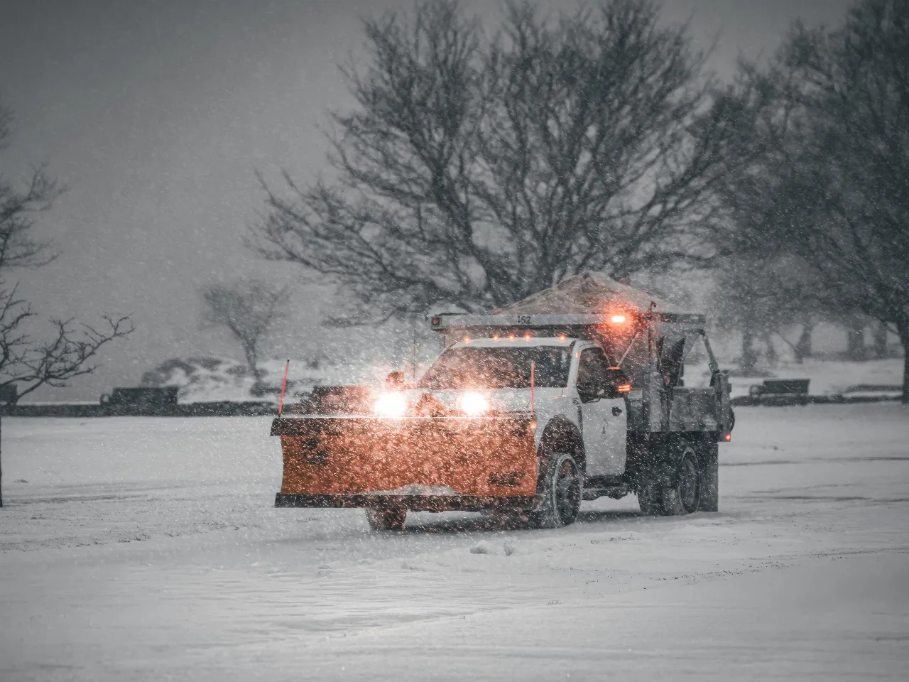 Un camion à lame orange dégage une route pendant qu'il neige, entre des arbres nus.