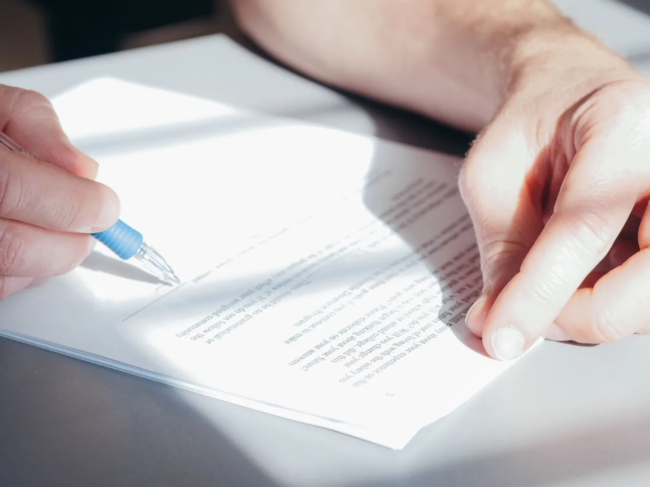 Deux mains signent un document sur une table de bois, au soleil rasant.