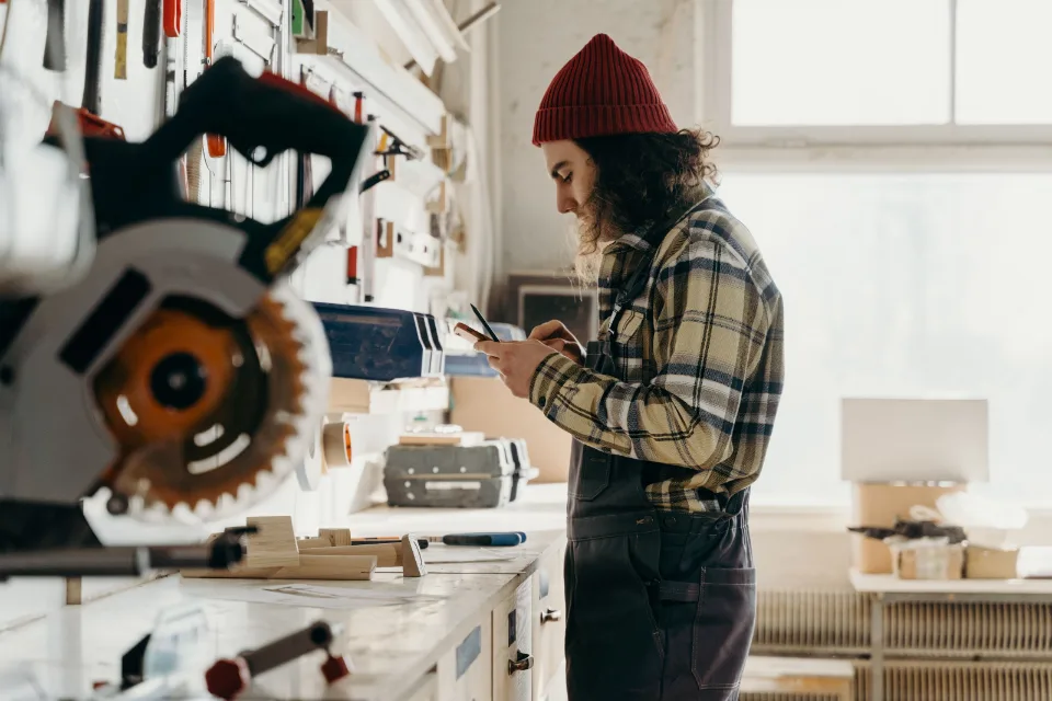 Un atelier de menuiserie : mur d'outils, scie a onglets, un telephone tenu en main devant l'etabli.