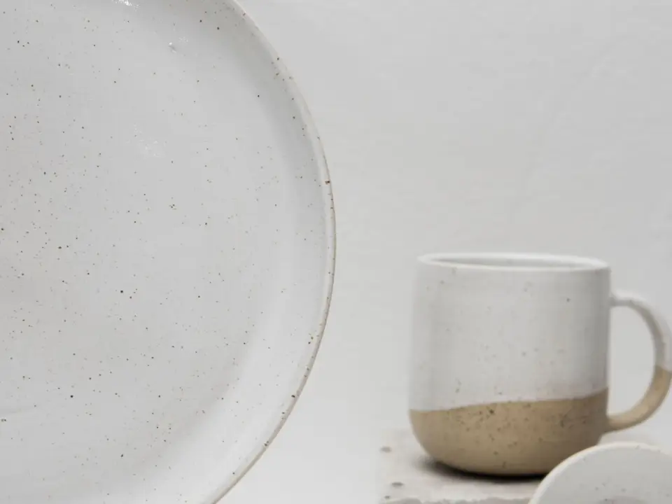 Une grande assiette de grès émaillé blanc moucheté, coupée par le bord du cadre, et une tasse à deux tons posée derrière, sur un fond clair.