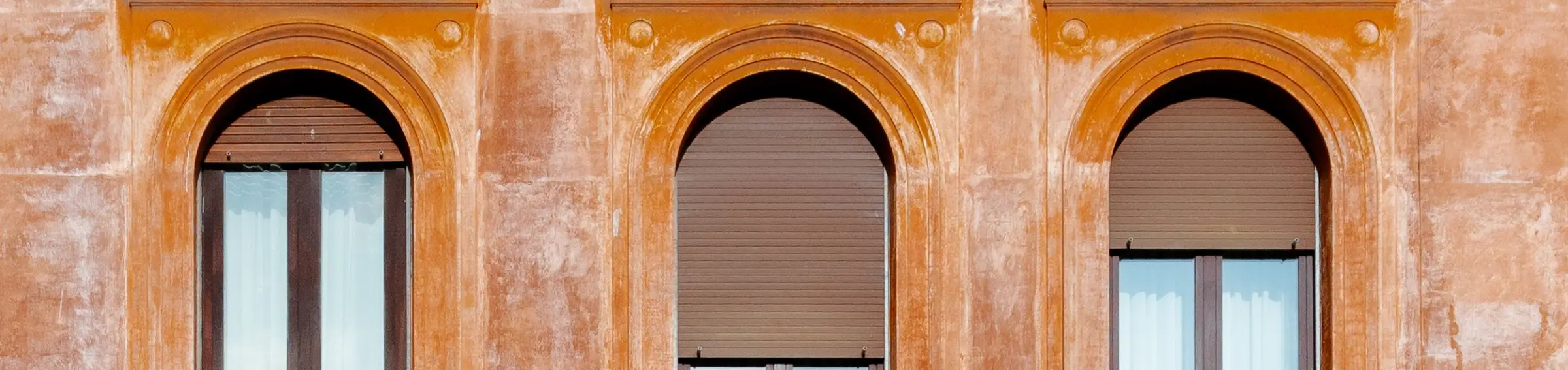 Façade d’un immeuble ancien, vue de face : trois fenêtres cintrées à corniche sous un enduit ocre lavé, persiennes baissées et rideaux clairs.