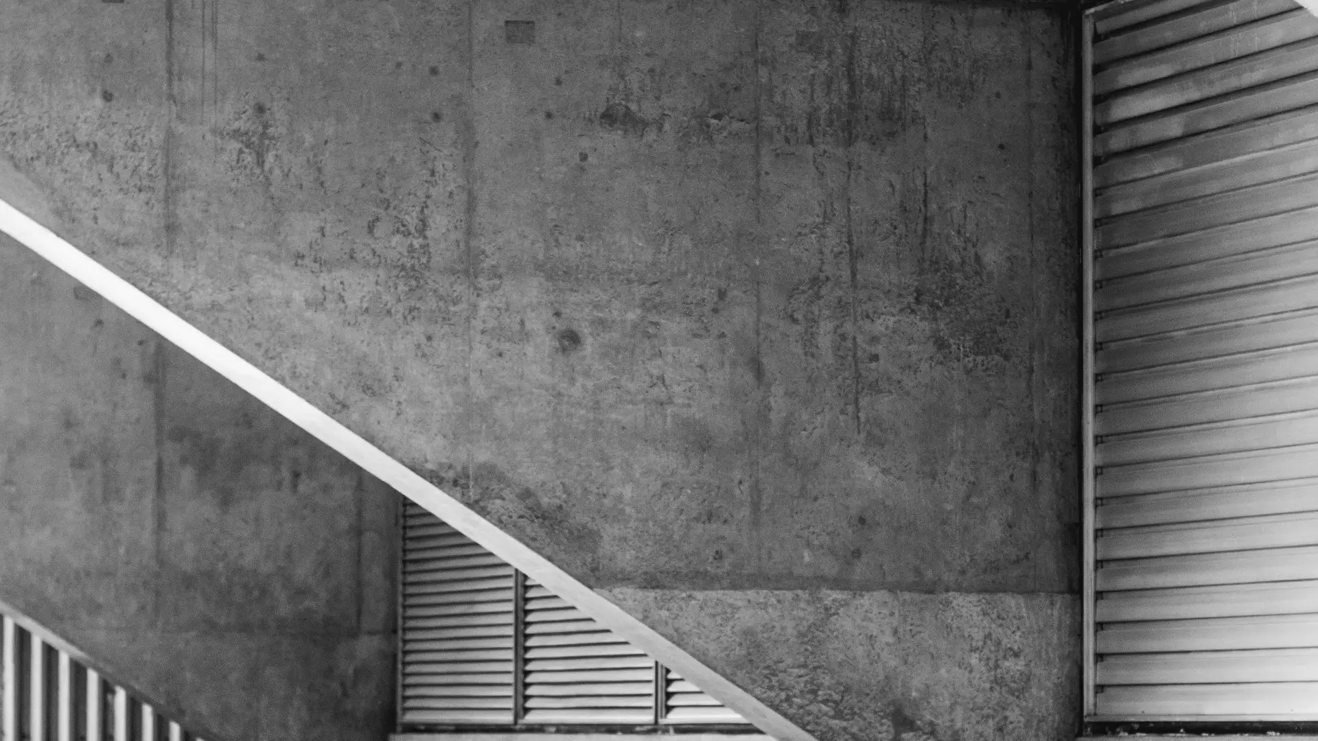 Sous-face de dalle et rampe d'escalier en diagonale sur un mur de béton, à côté d'un rideau de lamelles.