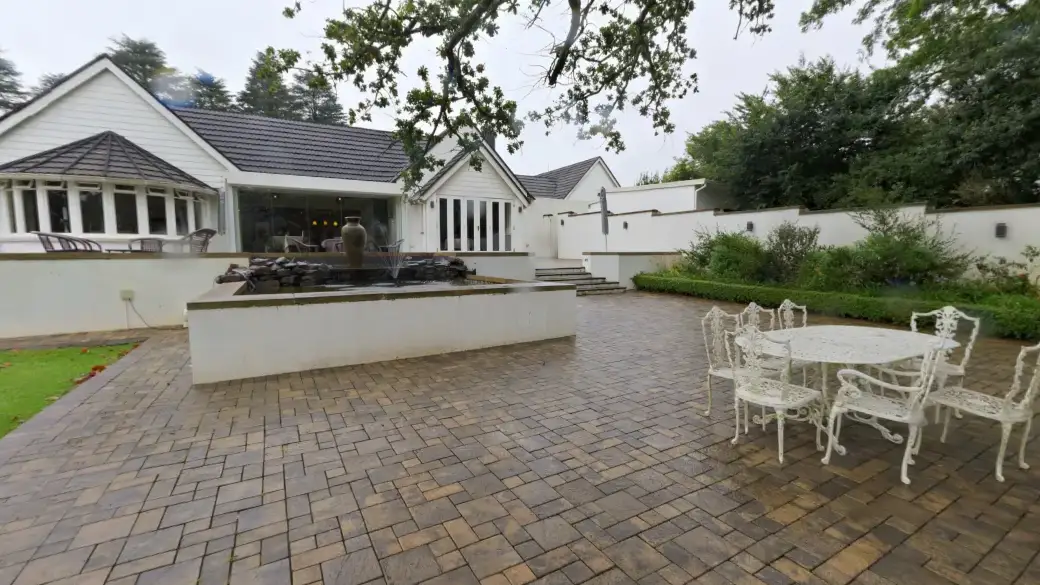 Terrasse pavée d'une maison de campagne blanche : un bassin, une table et des chaises en fer forgé, une grande baie vitrée ouvrant sur le salon.